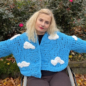 Op de afbeelding: Een blauwe gebreide cardigan met witte wolkenapplicaties. De cardigan is open aan de voorkant en heeft lange mouwen. Het model draagt de cardigan over een zwart shirt en een zwarte broek.