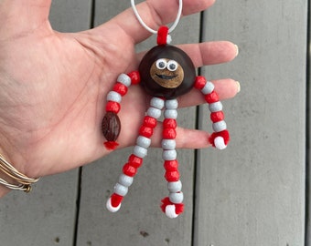 May include: A chestnut with googly eyes and a smiling mouth is strung with red and white beads to create a whimsical ornament.