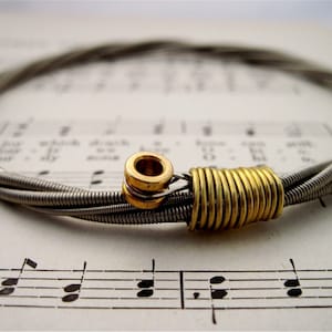 Può includere: Un braccialetto di corde di chitarra argentate con una chiusura in metallo dorato. Il braccialetto è su un foglio di musica.