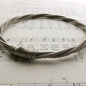 Può includere: Un braccialetto in corda di chitarra argentato con più fili di corda intrecciati insieme per formare un cerchio. Il braccialetto è su un foglio di musica.