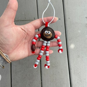 May include: A chestnut with a painted face and googly eyes is strung with red and gray beads to create a whimsical ornament.