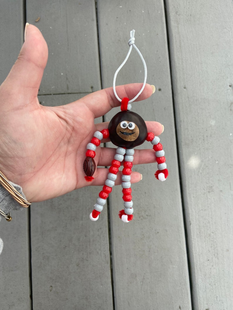 May include: A chestnut with a painted face and googly eyes is strung with red and gray beads to create a whimsical ornament.