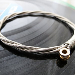 Può includere: Un braccialetto in corda di chitarra argentata con una chiusura dorata. Il braccialetto è realizzato con tre corde di chitarra intrecciate.
