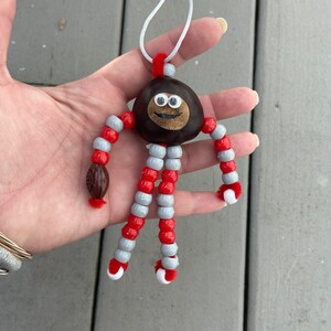 May include: A chestnut with googly eyes and a smiling mouth is strung with red and gray beads to create a whimsical figurine. A small brown football is attached to one of the bead legs.