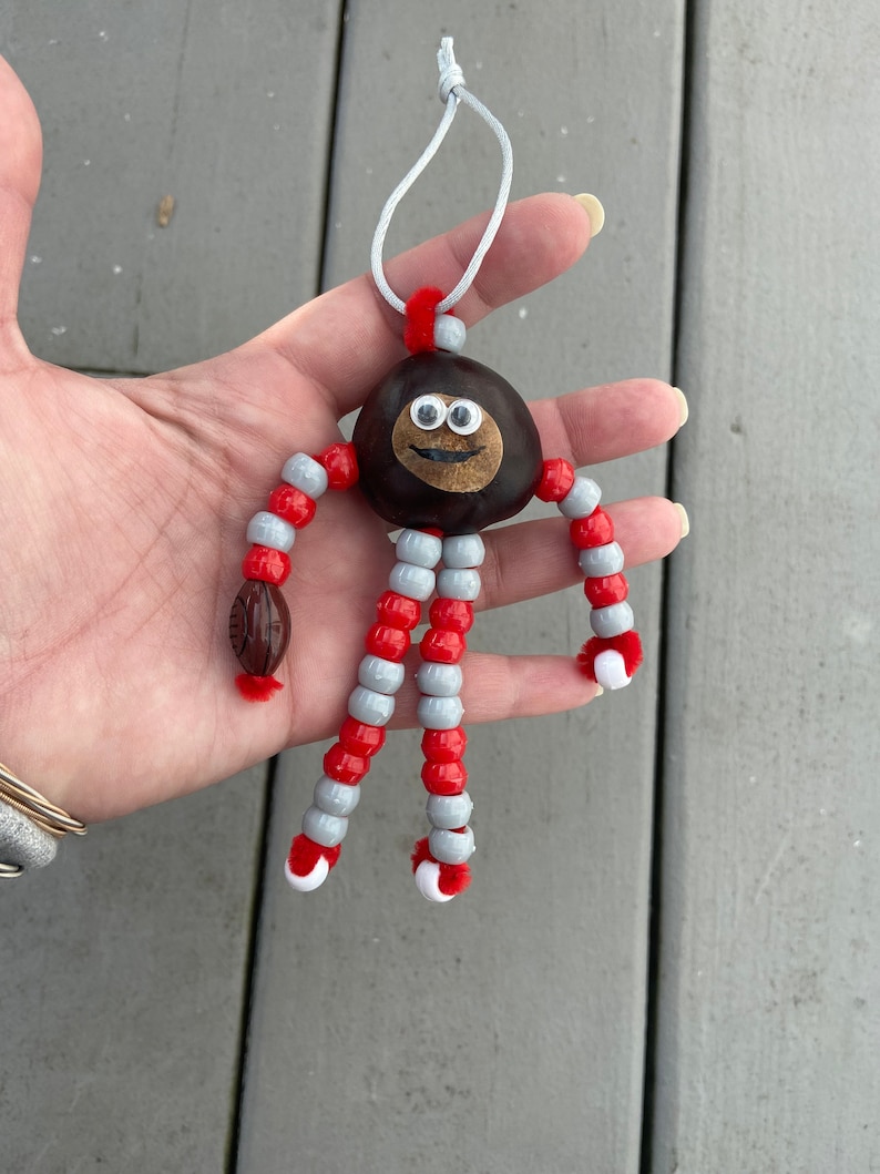 May include: A chestnut with googly eyes and a smiling mouth is strung with red and gray beads to create a whimsical figurine. A small brown football is attached to one of the bead legs.