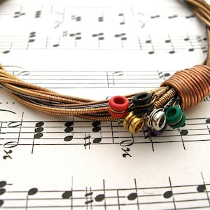 May include: A necklace made from multiple guitar strings with a copper wire clasp. The necklace has colorful beads attached to the ends of the strings.