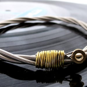 Op de afbeelding: Een zilveren gitaarsnaar armband met een gouden metalen sluiting. De armband is gemaakt van vier gitaarsnaren die samen gevlochten zijn.