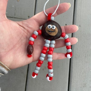 May include: A handmade ornament featuring a chestnut with googly eyes and a painted smile. The chestnut is attached to red and gray beaded limbs with white pom-poms at the ends.