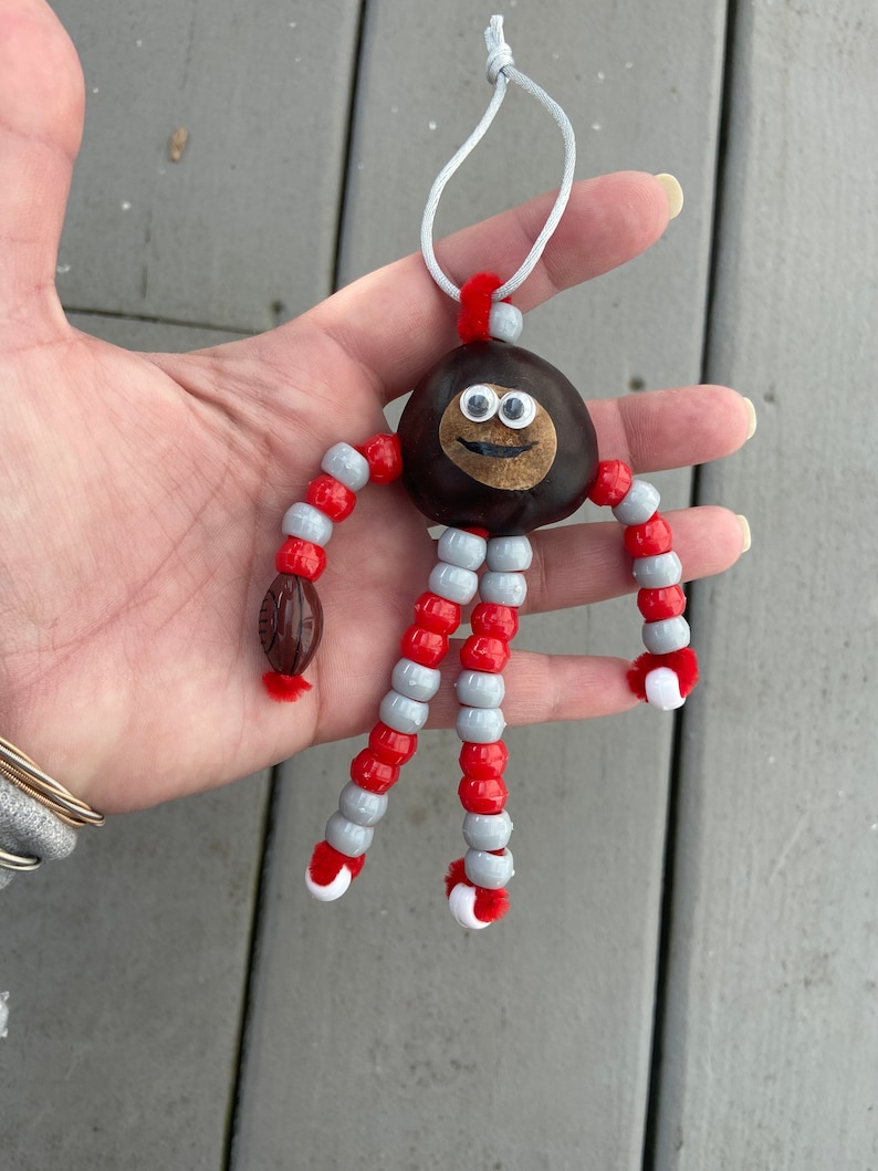 May include: A handmade ornament featuring a chestnut with googly eyes and a painted smile. The chestnut is attached to red and gray beaded limbs with white pom-poms at the ends.