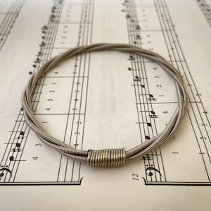 May include: A silver guitar string bracelet made from three guitar strings twisted together. The bracelet is lying on a sheet of music.