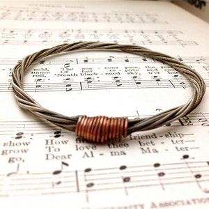 Peut inclure: Un bracelet en cordes de guitare argenté avec un accent en cuivre. Le bracelet est posé sur une feuille de musique.