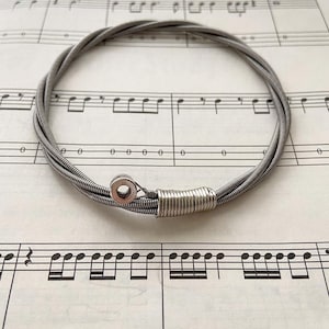 Può includere: Un braccialetto in corda di chitarra argentato realizzato con più corde di chitarra. Il braccialetto è su un foglio di musica.