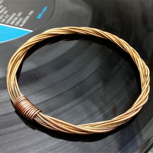 Könnte beinhalten: Ein Armband aus goldenen Gitarrensaiten mit geflochtenem Design. Das Armband liegt auf einer schwarzen Schallplatte.
