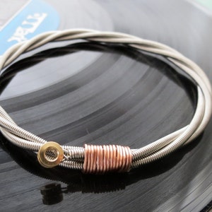 Può includere: Un braccialetto in corda di chitarra argentata con un accento in rame. Il braccialetto è su un disco in vinile nero.