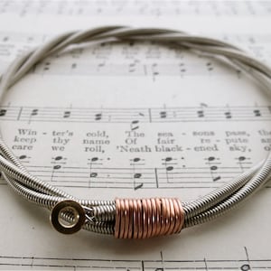 Può includere: Un braccialetto in corda di chitarra argentata con un accento in rame. Il braccialetto è realizzato con tre corde di chitarra intrecciate insieme. L'accento in rame è una piccola bobina di filo di rame avvolta attorno alle corde.