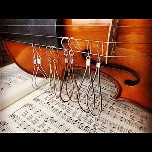 May include: Five pairs of earrings hanging on a violin. The earrings are made of silver and gold metal and have a teardrop shape. The violin is brown and has a natural wood finish.