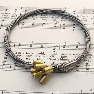 May include: A silver guitar string bracelet with a cluster of gold metal beads. The bracelet is lying on a sheet of music.