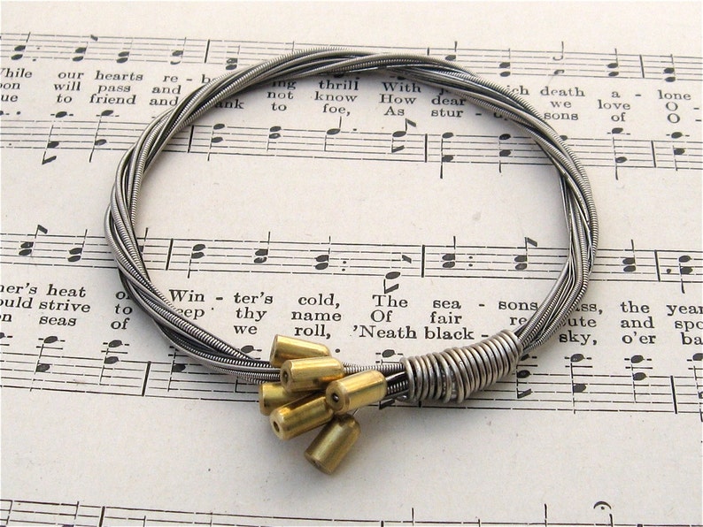 May include: A silver guitar string bracelet with a cluster of gold metal beads. The bracelet is lying on a sheet of music.