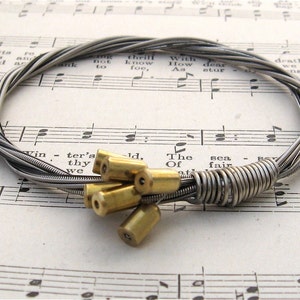 May include: A silver guitar string bracelet with a clasp made of multiple brass beads. The bracelet is lying on a sheet of music.