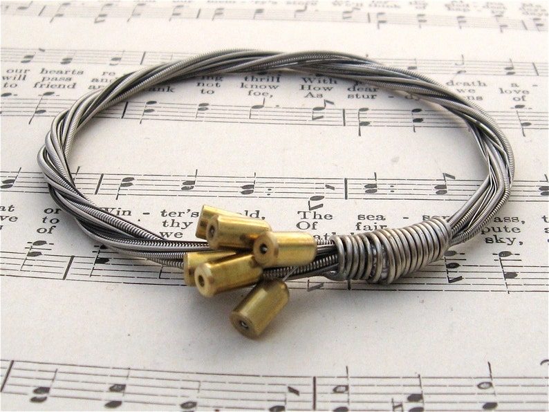 May include: A silver guitar string bracelet with a clasp made of multiple brass beads. The bracelet is lying on a sheet of music.
