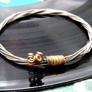 May include: A silver guitar string bracelet with a gold clasp. The bracelet is made from multiple guitar strings that are woven together.