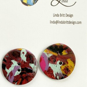 May include: Two colorful round buttons with a swirling abstract design in shades of red, yellow, black, white, pink, and green. The buttons have two holes each.