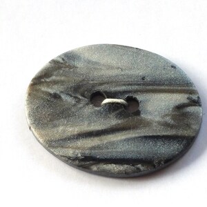 May include: Oval-shaped button with a marbled gray and black design. The button has two holes for sewing and a slightly irregular edge. The button is made of a shiny material.