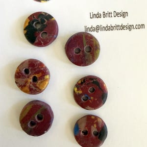 May include: Six round, two-hole buttons featuring a marbled pattern of deep red, yellow, black, and blue. The buttons are arranged on a white card printed with the text Linda Britt Design and the email address linda@lindabrittdesign.com.
