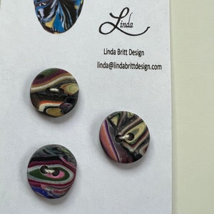 May include: Three colorful, round buttons with a swirl pattern. The buttons have two holes each. The buttons are on a white background with text that reads "Buttons as Individual as You!" and "Linda Britt Design" with an email address.
