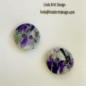 May include: Two round, two-hole buttons featuring a marbled pattern of deep purple, white, and muted green tones. The text Linda Britt Design and linda@lindabrittdesign.com is printed in black above the buttons on the white display card.