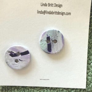 May include: Two round buttons with a marbled white, light purple, and dark purple pattern, each featuring two holes. The buttons are attached to a white display card printed with the text Linda Britt Design and linda@lindabrittdesign.com.