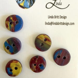 May include: Six round, two-hole buttons featuring a marbled pattern of blue, yellow, and deep red colors. The buttons are displayed on a white card printed with the text Linda, Linda Britt Design, and the email address linda@lindabrittdesign.com.