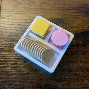 Pode incluir: Uma bandeja branca dividida com um quadrado amarelo, um círculo rosa e uma fileira de biscoitos em forma de coração. A bandeja está em uma superfície de madeira. O quadrado amarelo parece ser queijo, e o círculo rosa parece ser uma fatia de carne.