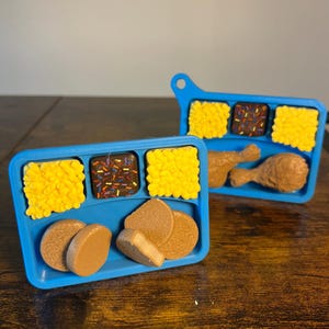 以下が含まれることがあります： おもちゃの食べ物が入った2つの青いトレイ。1つのトレイには黄色の正方形、スプリンクル付きの茶色の正方形、茶色の円があります。もう1つのトレイには、黄色の正方形、スプリンクル付きの茶色の正方形、2つの茶色のドラムスティック、茶色のボールがあります。
