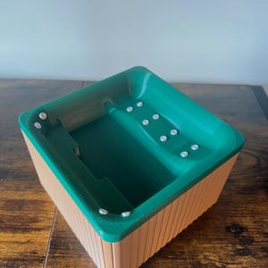 Könnte beinhalten: Ein Miniatur-Whirlpool-Modell mit grünem Innenraum und beigefarbenem Äußeren. Der Whirlpool hat eine rechteckige Form mit abgerundeten Ecken und kleinen, weißen, kreisförmigen Details. Das Äußere hat eine vertikale Holzmaserung.