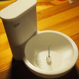 May include: White plastic toilet-shaped bowl with a small screw in the center. The bowl is designed to hold small items.