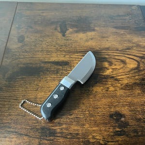 Könnte beinhalten: Ein kleines Messer aus Edelstahl mit einem schwarzen Griff und drei silbernen Nieten. Das Messer hat eine gebogene Klinge und eine silberne Kette am Griff. Das Messer liegt auf einer dunkelbraunen Holzoberfläche.