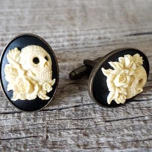 May include: A pair of cufflinks with a black background and a white cameo of a skull with flowers.