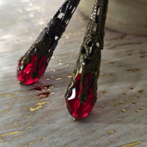 May include: Two ornate, dark metal hair sticks with a teardrop-shaped, faceted red crystal at the end. The metalwork features intricate cut-out designs. The background is a light, textured surface with gold accents.