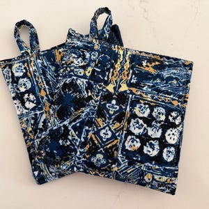 May include: Two square pot holders with loop handles. The pot holders have a repeating pattern of white, gold, and black shapes on a navy blue background. The pot holders are made of a quilted fabric.