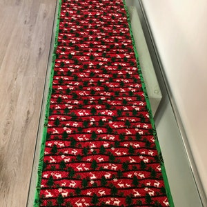 May include: A red and green Christmas table runner with a white reindeer and tree pattern. The runner has a green border.