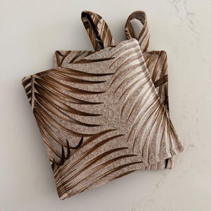 May include: Two square pot holders with loop handles. The pot holders feature a repeating pattern of brown palm fronds on a beige background. The edges are neatly sewn, and the loop handles are made of the same fabric.