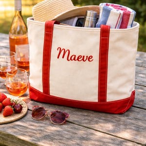 May include: Cream-colored tote bag with red accents, embroidered with the name "Maeve." The bag is filled with a straw hat, towel, and a stainless steel water bottle. Sunglasses and a plate of strawberries are on the wooden table.