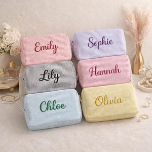 May include: Six personalized cosmetic bags in various colors, including pink, black, blue, and yellow. Each bag features a different name embroidered in a complementary color. The bags have a striped pattern.