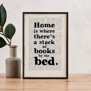 May include: A black and white print with the text "Home is where there's a stack of books by the bed." The print is framed in a black frame and is displayed against a white wall.
