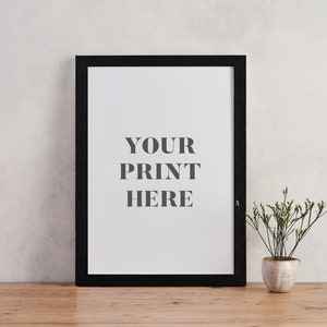 May include: A black picture frame with a white mat and the text "YOUR PRINT HERE" on a white background. The frame is sitting on a wooden table next to a small potted plant.