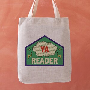 May include: A light gray tote bag with cream-colored handles. The bag features a graphic with the text "YA READER" in white and red, set against a green cloud and lightning bolts, within a purple and green house-shaped border.