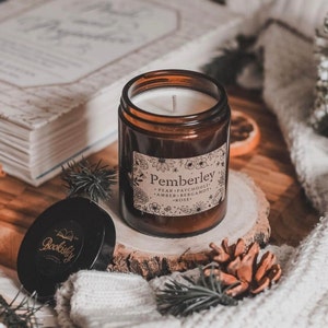May include: A brown glass candle jar with a white candle and a label that reads "Pemberley". The label lists the scents: pear, patchouli, amber, bergamot, and rose. The candle sits on a wooden slice with a pine cone and a lid that reads "Bookishly".