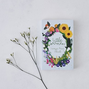 May include: A book cover featuring a floral wreath design with sunflowers, oranges, and various flowers in shades of pink, purple, and blue. The title "Little Women" is written in green text, and the author "Louisa May Alcott" is written in black text.