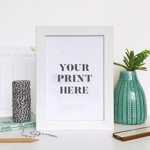 May include: White picture frame with a white mat and a white background. The text "YOUR PRINT HERE" is printed in black on the white background. The frame is sitting on a white surface with a spool of twine, two books, a pencil, a vase with a green plant, and a brown object.